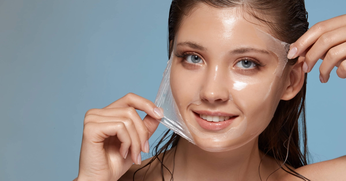 Smiling woman with wet hair peeling a clear facial mask (peel-off) from her moist, shiny face, related to Chemical Peels in Minot, ND. Light blue background.